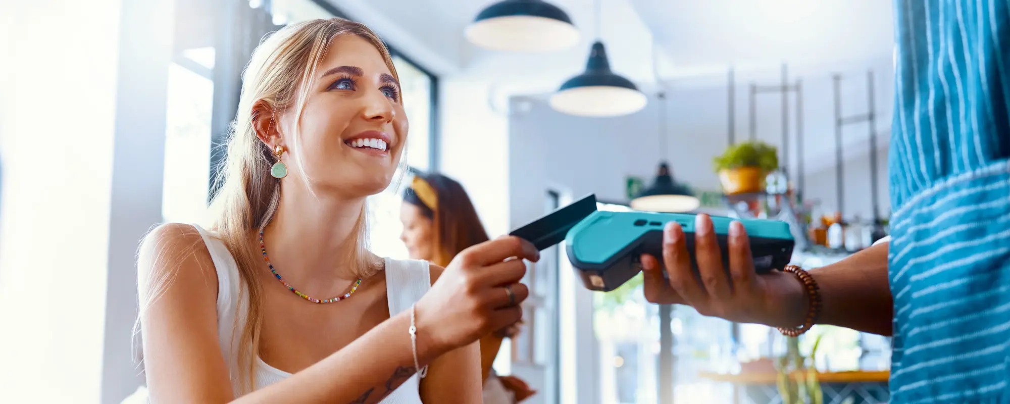 A young woman sits in a kitchen smiling and holding her debit card out to pay.